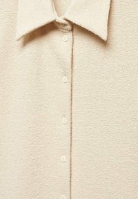 Chemise beige à boutons, fabriquée en tissu texturé, avec un col étalé et six boutons ronds blancs sur la patte de boutonnage avant.