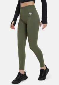 Olivi vihreät korkeavyötäröiset legginsit, joissa on sileä pinta ja valkoinen logo reidessä. Yhdistetty mustiin urheilukenkäisiin.