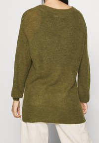 Personne portant un pull tricoté ample vert olive et un pantalon crème, vue de dos sur un fond clair uni.