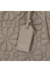 Tessuto beige trapuntato con cuciture a forma di cuore; presenta un'etichetta rettangolare con "kate spade" inciso in oro, attaccata da una sottile cinghia.