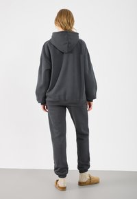 Sudadera gris oscuro con una gran capucha ajustable, combinada con pantalones de chándal a juego. Tejido cómodo, corte relajado, puños y bajos acanalados.