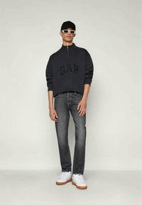 Sweatshirt preto com fecho e logotipo "GAP", combinado com jeans escuros e ténis brancos. Os acessórios incluem um boné preto e óculos de sol brancos.
