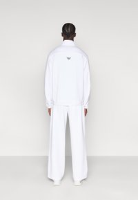 Λευκή φόρμα που περιλαμβάνει ζακέτα με φερμουάρ και ελαστικές μανσέτες, καθώς και φαρδιά παντελόνια. Διακριτική λεπτομέρεια με λογότυπο στην πλάτη της ζακέτας.