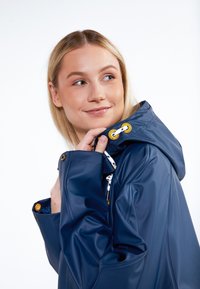 Vízálló sötétkék dzseki, állítható kapucnival, arany színű fém kiegészítőkkel és texturált felülettel. Oldalsó zsebekkel és díszítő varrással rendelkezik.