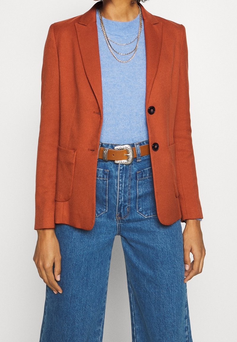 Roestoranje blazer met ingesneden revers, twee knopen en voorzakken, gedragen over een lichtblauwe trui, gecombineerd met een high-waisted spijkerbroek.