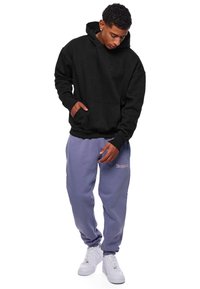 Černá mikina s vyraženým logem, kombinována se světle fialovými tepláky s výšivkou značky. Bílé tenisky doplňují outfit.