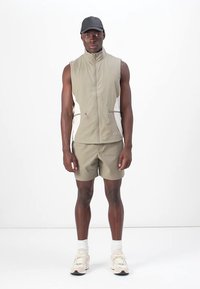 Lightweight, beige sleeveless vest with front zipper and side pockets paired with matching shorts. Worn with a black cap and sneakers.