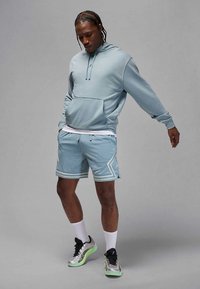 Ljusblå munkjacka med huva och en framficka, tillsammans med matchande meshtights med vita geometriska mönster, kombinerade med silverfärgade och gröna sneakers.