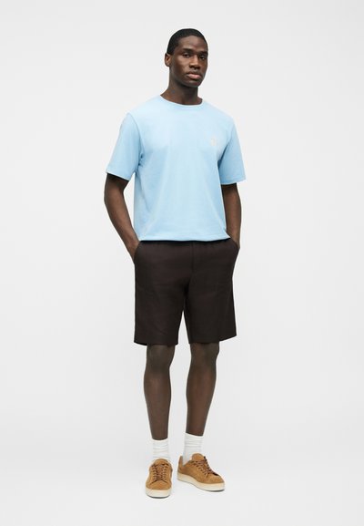 Homme debout, les mains dans les poches, portant un t-shirt bleu clair, un short marron foncé, des chaussettes blanches et des baskets en daim beige sur fond blanc.