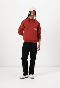 Rød hettegenser med "POMPE" tekst på brystet, svarte bukser og hvite sneakers med grå detaljer; iført en lys beige caps.