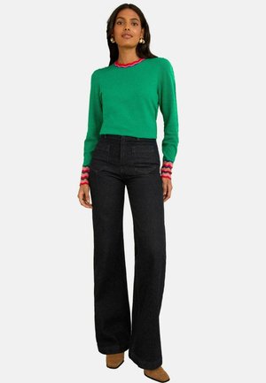 Pull en tricot vert avec un col et des poignets en forme de coquille rouge et rose ; associé à un jean en denim sombre taille haute, à jambes larges et à des bottines brunes.