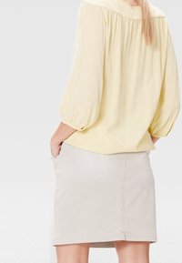 Vrouw met lang haar in een lichtgele blouse met pofmouwen en een beige knie-lange rok, handen in de zakken, met haar rug naar de kijker gekeerd.