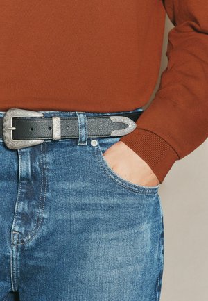 Person wearing blue jeans with a black leather belt featuring silver buckle accents, hand resting in front pocket, rust-colored long sleeve top.