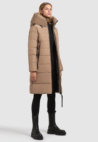 Beige puffjakke med stor hætte, lynlås lukning og side lommer. Har quiltet design og tekstureret stof. Bæres med sorte leggings og støvler.