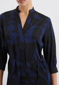Robe chemise boutonnée en noir avec un motif abstrait bleu profond, en tissu léger, col montant et ceinture à la taille. Manches trois-quarts.