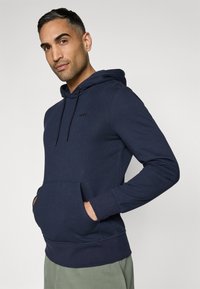 Corta-vento azul marinho feito de tecido macio, apresentando um bolso canguru frontal, capuz com cordão e um logotipo discreto no peito.
