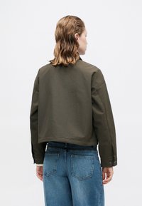 Olijfgroene cropped jack met een ontspannen pasvorm en brede mouwen, voorzien van een met knopen sluitende manchette. Gecombineerd met blauw hoge taille denim jeans.