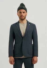 Sacou bleumarin din țesătură netedă, cu două buzunare frontale și închidere cu nasture. Purta peste un tricou bej deschis, asortat cu pantaloni închiși la culoare.