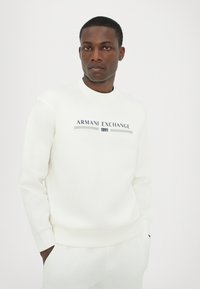 Fehér kapucnis pulóver az "Armani Exchange 1991" logóval sötétkék és szürke színben, kerek nyakkal és bordázott mandzsettával. Sima textúra, laza dizájn.