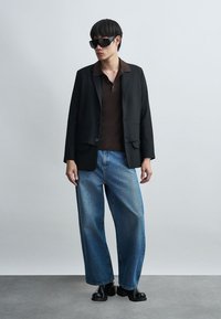 Άνδρας που φοράει υπερμεγέθες μαύρο σακάκι, σκούρο καφέ πόλο, μπλε τζιν παντελόνι με φαρδιά μπατζάκια, μαύρες μπότες και μεγάλα μαύρα γυαλιά ηλίου μπροστά σε απλό τοίχο.