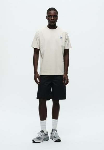 T-shirt en coton beige clair avec un logo bleu, associé à un short noir. Porté avec des baskets grises et des chaussettes blanches, se tenant devant un fond uni.