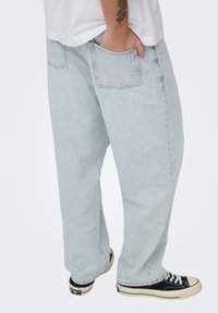 Φωτεινό γαλάζιο τζιν με φαρδιά εφαρμογή, με πίσω τσέπες και ξεφτισμένο τελείωμα, σε συνδυασμό με μαύρα και λευκά αθλητικά παπούτσια χαμηλού ύψους.