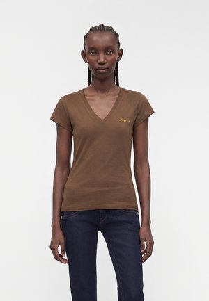 Vrouw met gevlochten haar die een bruin T-shirt met V-hals en korte mouwen en donkerblauwe spijkerbroek draagt, staand tegen een effen witte achtergrond.