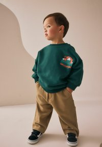 Sweatshirt vert avec un graphisme "Aventure en plein air", pantalon beige ample et baskets noires, sur un fond clair.