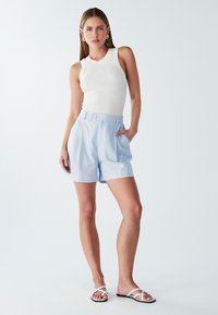 Světle modré kraťasy s vysokým pasem, na kterých jsou skladby a detail zapínání na knoflík vpředu, spárované s bílým žebrovaným tílkem a bílými sandály na řemínky.