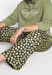 Vihreä huppari, jossa on brodeerattu logo, yhdistetty mustiin housuihin, joissa on keltainen kukkakuosi. Kangas näyttää pehmeältä ja sillä on rento istuvuus.