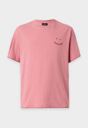 Homme portant une chemise rose à manches courtes, un jean bleu et des mocassins jaunes, debout avec une main dans la poche devant un fond uni.