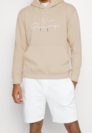 Sweat à capuche beige avec poche avant et inscription blanche « New Beginnings », porté avec un short blanc longueur genou sur une personne debout.