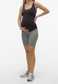 Zwarte, aansluitende tanktop gecombineerd met lichtgrijze, aansluitende shorts. Grijze sneakers met witte accenten. Vrouw staat met één hand op haar buik.