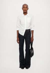 Hvitt langermet skjorte med brystlomme, kombinert med mørke flared jeans med knappedetaljer ved mansjettene, og holder en svart håndveske.