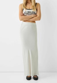 Žena se založenýma rukama, oblečená v bílém kratším tílku s nápisem, dlouhé bílé sukni a černých síťovaných balerínkách na bílém pozadí.