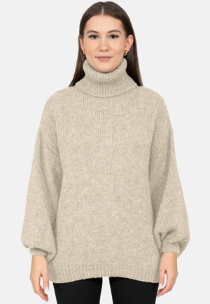 Vrouw draagt een beige gebreide coltrui met ballonmouwen en een zwarte broek, staand tegen een effen lichte achtergrond.