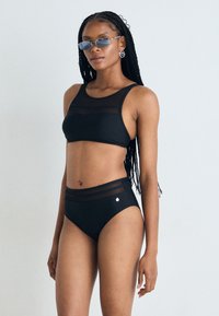 Juodas bikini rinkinys, turintis trumpą viršų su tinklinėmis detalėmis ir aukštu juosmeniu. Medžiaga yra lygi, o dizainas elegantiškas.