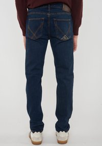 Persona che indossa jeans blu scuro con cuciture distintive sui taschini posteriori e sneakers bianche, in piedi su uno sfondo neutro.