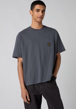 Joven con una camiseta de gran tamaño gris oscuro con un bolsillo en el pecho y pantalones negros, de pie frente a un fondo gris liso.
