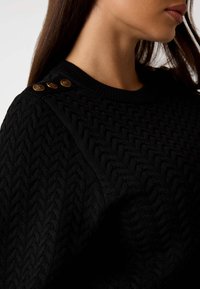 Maglione nero lavorato a maglia con motivo testurizzato, scollatura a girocollo a costine e tre bottoni decorativi dorati sulla spalla.