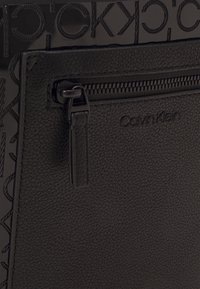 Zbliżenie na czarną, teksturowaną skórzaną saszetkę Calvin Klein z kieszenią na zamek i wytłoczonym logo na wzorzystym tle.