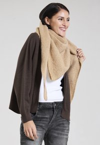 Beige scarf made of soft material draped over a brown cardigan, paired with a white top and gray jeans, showcasing a relaxed fit.