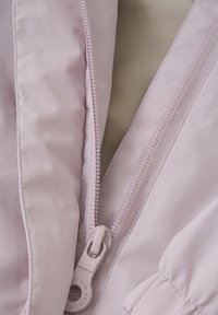 Lyserød jakke med en glat, tekstureret stof og en central lynlås med en rund trækknap. Syede detaljer er synlige.