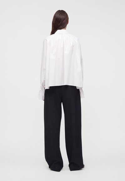 Femme aux longs cheveux bruns porte un chemisier blanc ample et un pantalon noir large, debout de dos devant un fond clair uni.