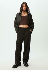 Sort læderjakke, brun crop top og brede sorte bukser. Mørkt fodtøj med tykke såler. Model står mod en lys baggrund.