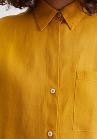 Mosterdgele linnen blouse met een button-down kraag, een voorste knoopsluiting en een enkele borstzak. Soepele textuur en lichtgewicht ontwerp.