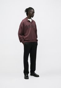 Ung mand med dreadlocks iført en vinrød HUGO-pullover, sorte bukser og sorte sko, der står mod en ensfarvet hvid baggrund.