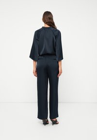Navy Satin Zweiteiler bestehend aus einem Langarm-Oberteil mit gewelltem Saum und einem weiten Hosen mit elastischem Bund. Sichtbare High Heels.
