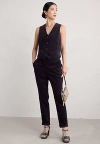 Temnozeleni žametni brezrokavnik s tremi gumbi, v kombinaciji s krojenimi hlačami. Model nosi čevlje z visokim peto in drži majhno torbico. Teksturni materiali.