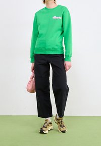 Grön sweatshirt med rosa logotyp, matchad med svarta vida byxor och beige sportskor. Bär en liten rosa väska. Enkel bakgrund.